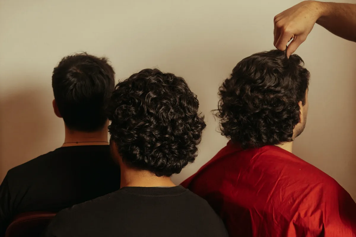 تلات شباب قاعدين في كراسي حلاقة من ورا، واحد شعره ناعم مفرود والتاني مويفي والتالت كيرلي، وإيد الحلاق بترفع خصلة تقارن بينهم