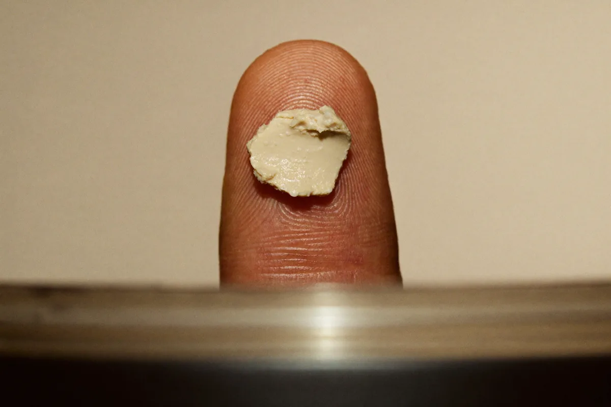 Extreme close-up of a fingertip holding a fingertip-sized dab of matte hair wax against a pale background